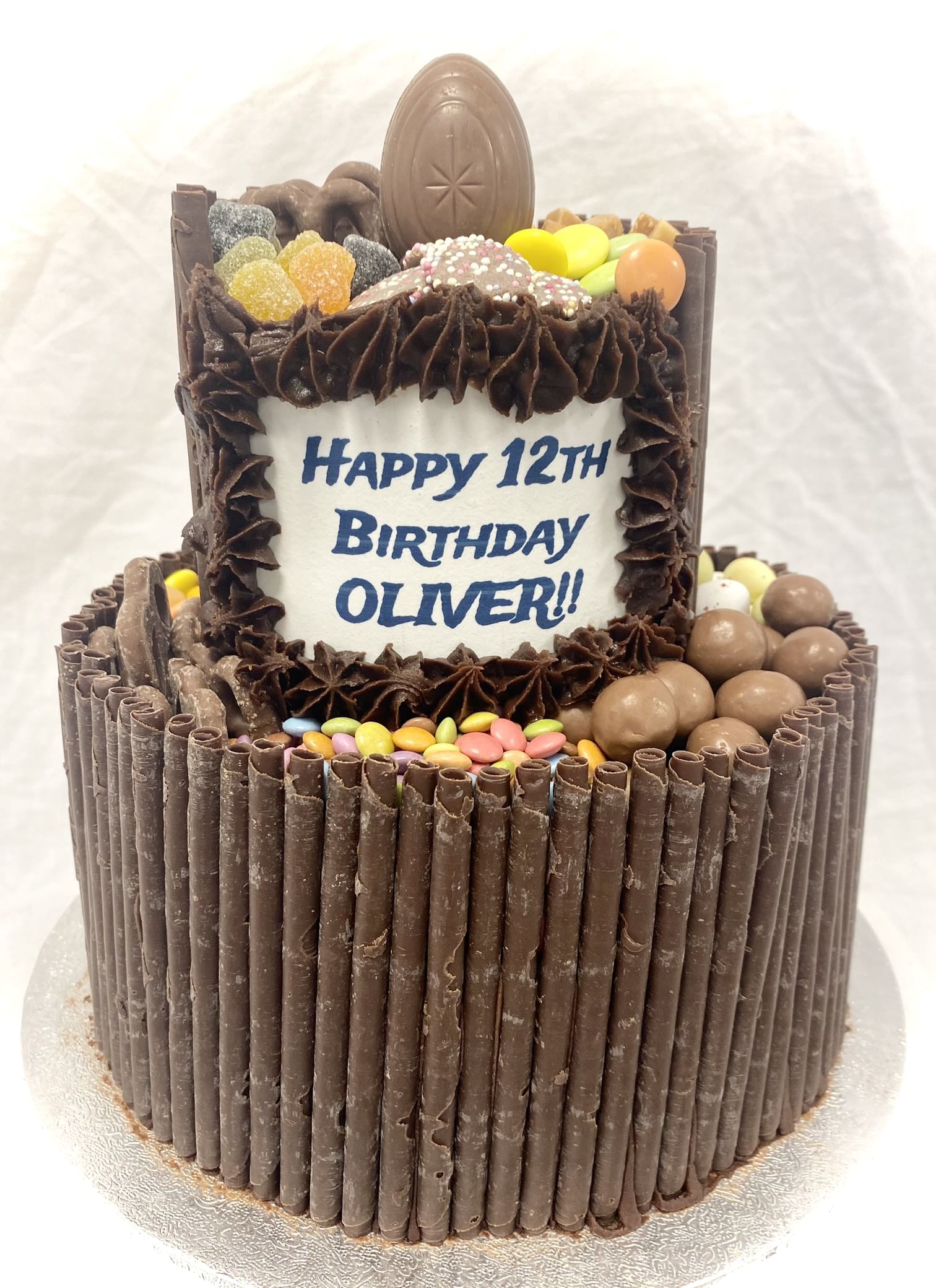 A two-tier Chocolate Blackout Cake decorated with various chocolates and colorful candies, featuring a rectangular sign on the front that reads 'Happy 12th Birthday Oliver!!' in blue lettering. An oval chocolate sits at the top center, and the sides are lined with chocolate sticks over chocolate frosting.